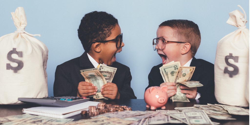 Twee kinderen in pak lachen en tellen geld, met zakken vol dollarbiljetten en een spaarvarken op tafel, als metafoor voor financiële afwegingen bij het inschakelen van een makelaar