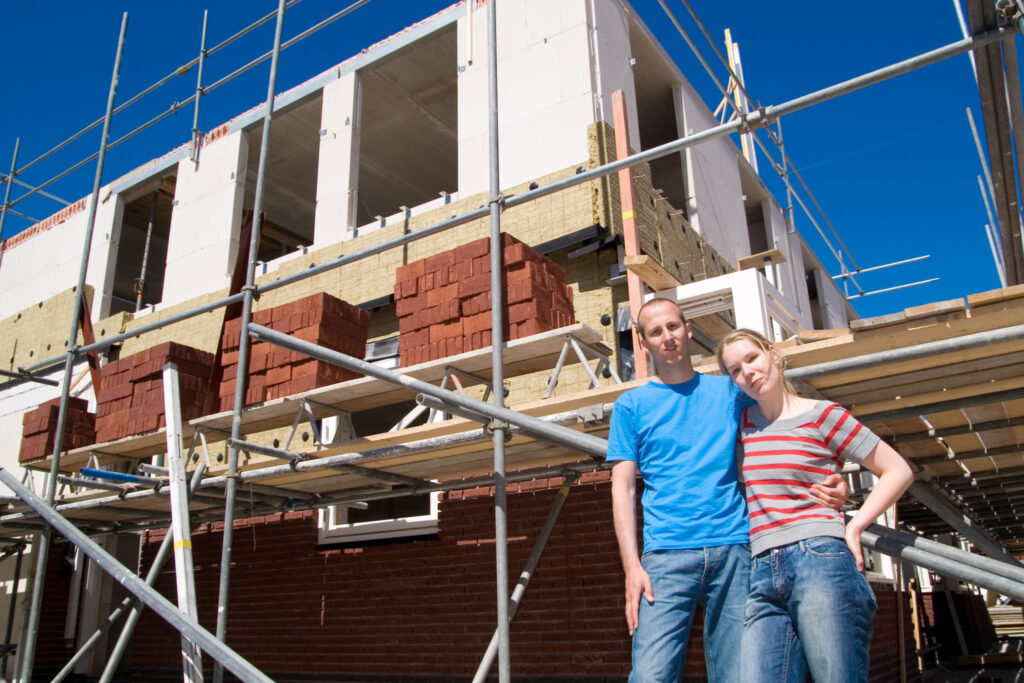 Jong stel staat voor een in aanbouw zijnde nieuwbouwwoning, omringd door steigers en bouwmaterialen. Dit beeld illustreert de uitdagingen én hoopvolle vooruitzichten van de woningmarkt in 2025.