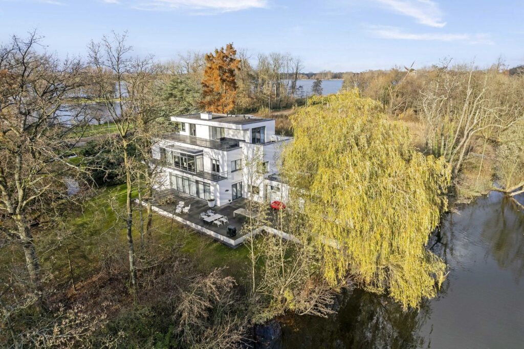 Unieke zijaanzicht van de iconische villa Plaszoom 1, omgeven door water en groen, met uitzicht op de Kralingse Plas en een groot terras dat naadloos overgaat in de natuur.