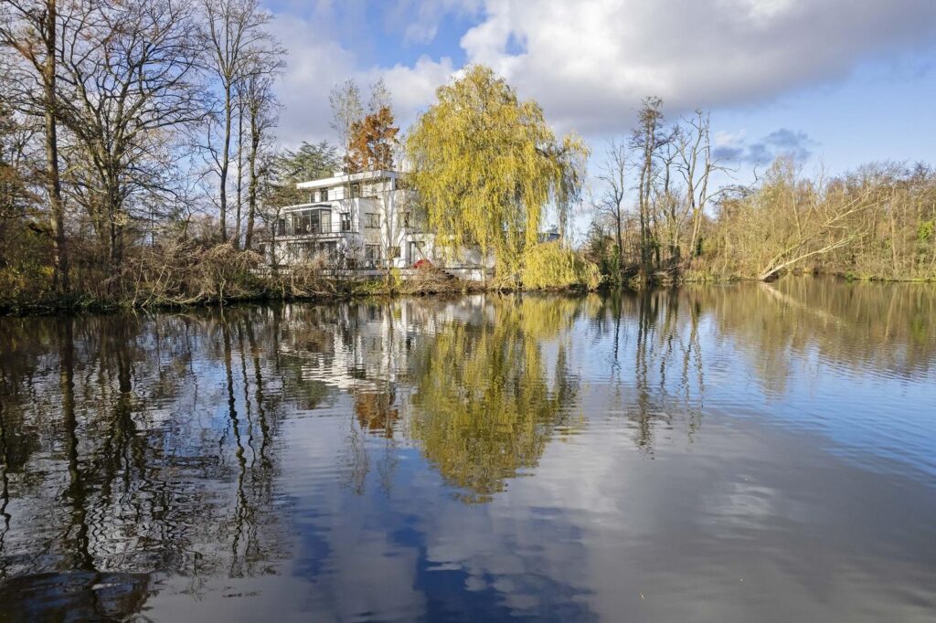 Villa Plaszoom 1 weerspiegeld in het water van het Kralingse Bos, omringd door volgroeide bomen en een serene natuurlijke omgeving.