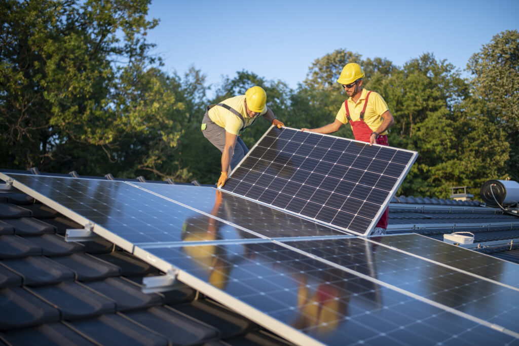 Twee monteurs installeren zonnepanelen op een dak, dragen gele helmen en veiligheidsuitrusting, duurzame energie-installatie in een groene omgeving.