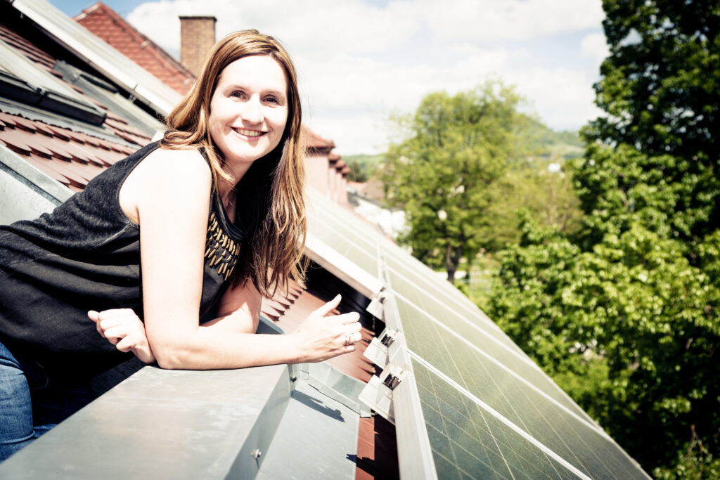 Vrouw leunt tegen balkon naast zonnepanelen op het dak, glimlachend en genietend van zonnig weer, duurzaam energiegebruik.