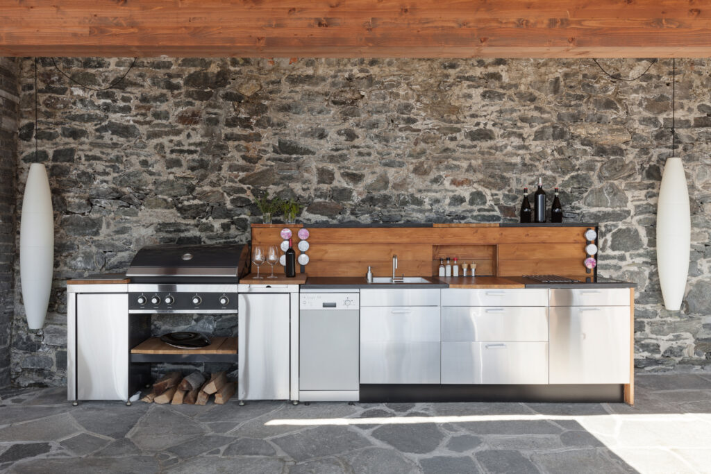 Moderne buitenkeuken met roestvrijstalen apparatuur en houten details, tegen een stenen muur.
