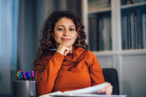 Vrouw in een oranje trui zit aan een bureau met documenten en een pen, klaar om financiële keuzes te overwegen voor de overstap van een huurwoning naar een koopwoning