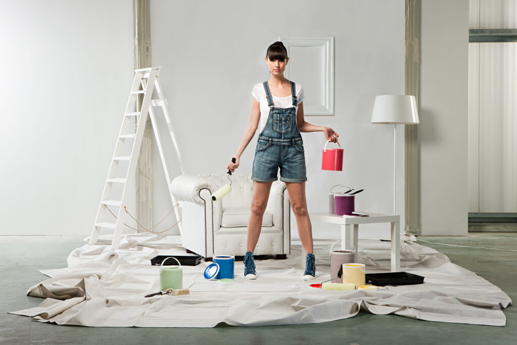 Vrouw in een ruimte bezig met schilderen, omringd door verfblikken en gereedschap – illustratie van een van de tips om de woningwaarde te verhogen door het huis op te frissen met een nieuwe verflaag