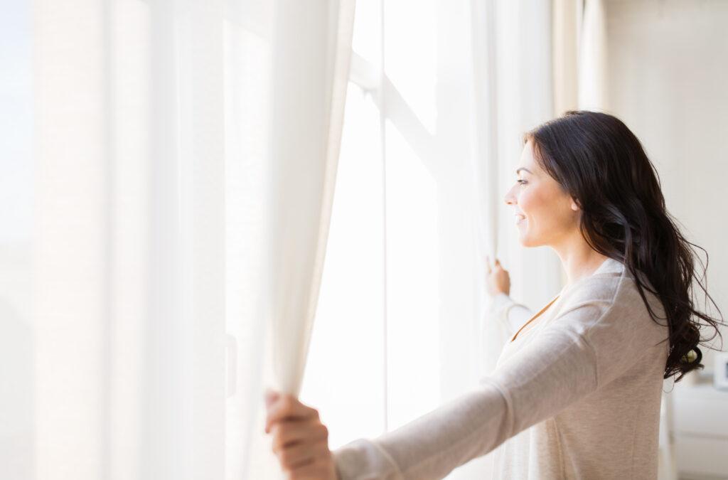Vrouw opent lichte gordijnen in een zonnige woonkamer en geniet van de lichtinval.