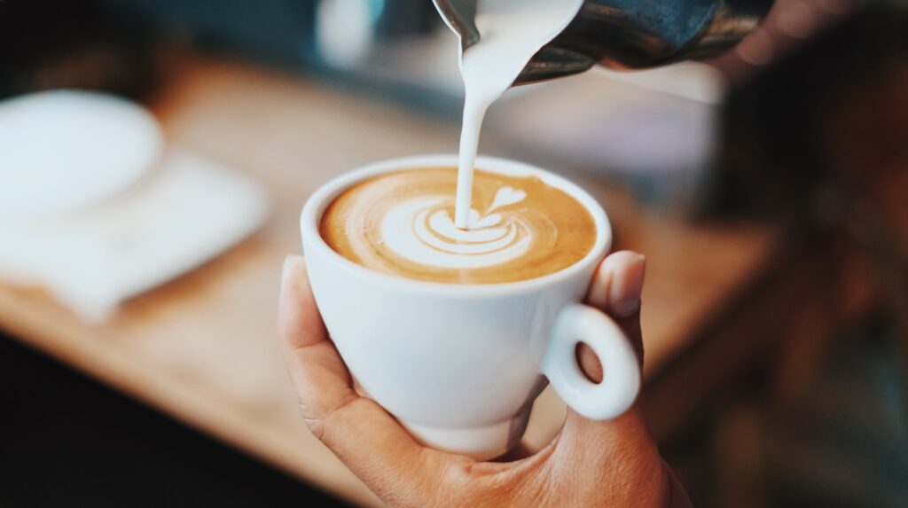 Close-up van een hand die een cappuccino vasthoudt terwijl er warme melk in wordt geschonken, met een prachtig latte art-patroon.