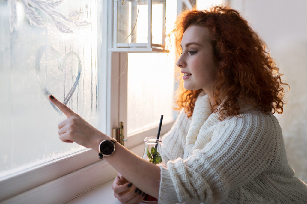 Vrouw tekent een hartje op een beslagen raam als symbool voor frisse lucht en het belang van ventilatie in huis.