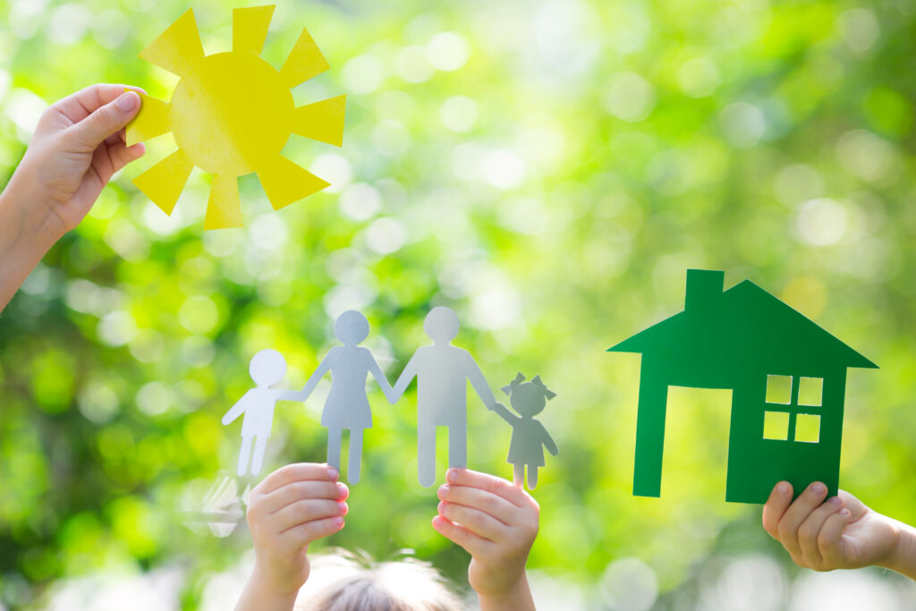 Papieren figuren van een gezin, een huis en een zon, vastgehouden tegen een achtergrond van groene natuur, als symbool voor duurzaam wonen en milieuvriendelijke keuzes