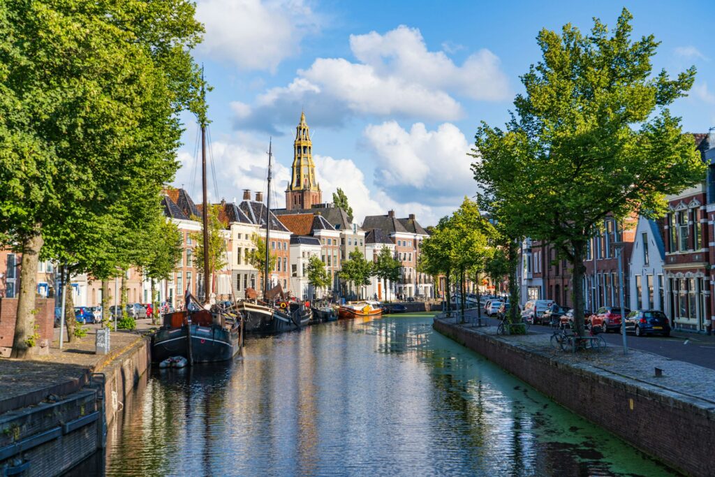 Gezicht op een historische gracht in Groningen met groene bomen, traditionele herenhuizen en boten aan het water. De Martinitoren steekt uit boven de stad. Dit beeld illustreert de woningmarkt in Groningen en de stijgende huizenprijzen in 2025