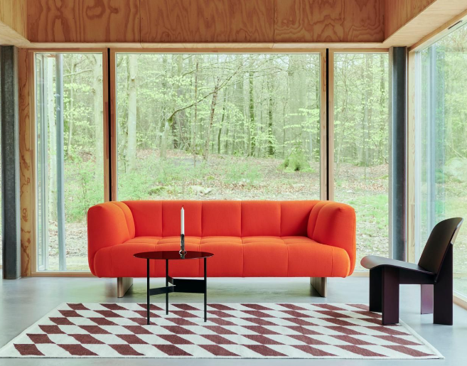 Moderne woonkamer met een opvallende oranje bank uit de HAY Voorjaarscollectie 2025, gecombineerd met een geometrisch vloerkleed en minimalistische meubels.