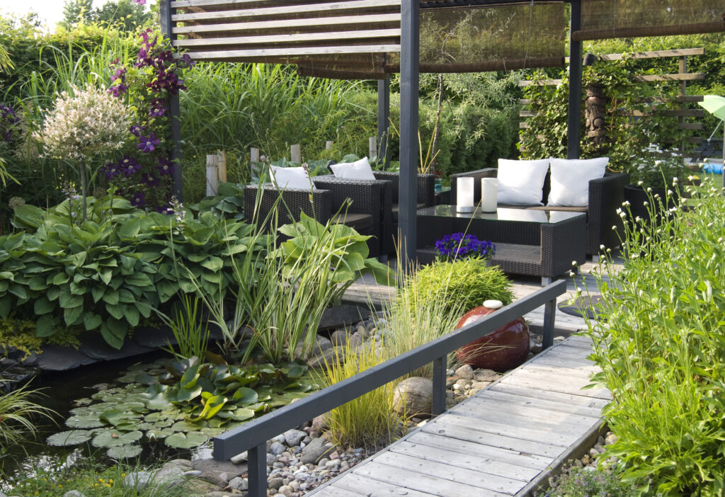 Sfeervolle groene tuin met vijver, houten vlonder en een moderne zithoek onder een pergola, omringd door weelderige beplanting.