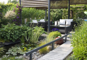 Sfeervolle groene tuin met vijver, houten vlonder en een moderne zithoek onder een pergola, omringd door weelderige beplanting.