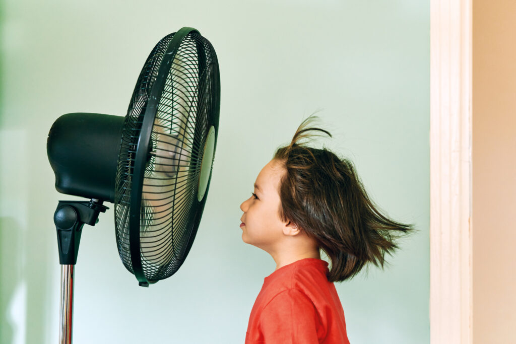Kind staat voor een ventilator, haar wappert in de luchtstroom, symbolisch voor frisse lucht en ventilatie in huis
