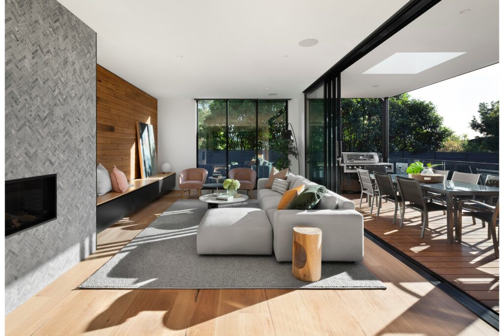 Modern ingerichte woonkamer met grote schuiframen en een naadloze overgang naar een zonnig terras, omringd door groen. Het interieur combineert natuurlijke materialen zoals hout en zachte stoffen voor een luxe en warme uitstraling.