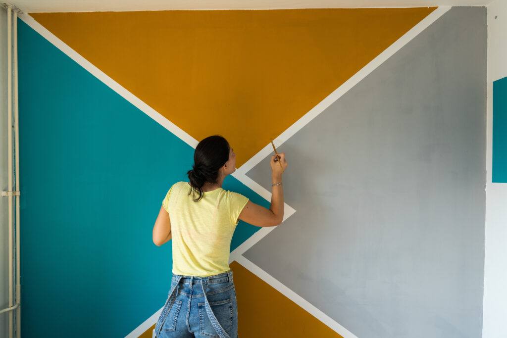 Een vrouw schildert geometrische kleurvlakken in okergeel, turquoise en grijs op een muur, passend bij de interieurtrend van kleurzones.