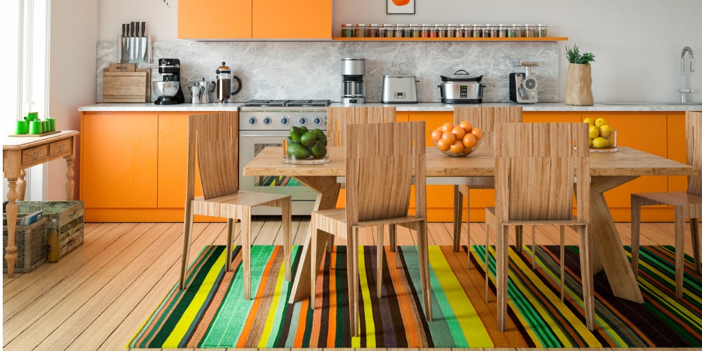 Eclectisch interieur met een mix van moderne en houten meubels, feloranje keukenkasten en een kleurrijk gestreept vloerkleed – woontrend 2025.