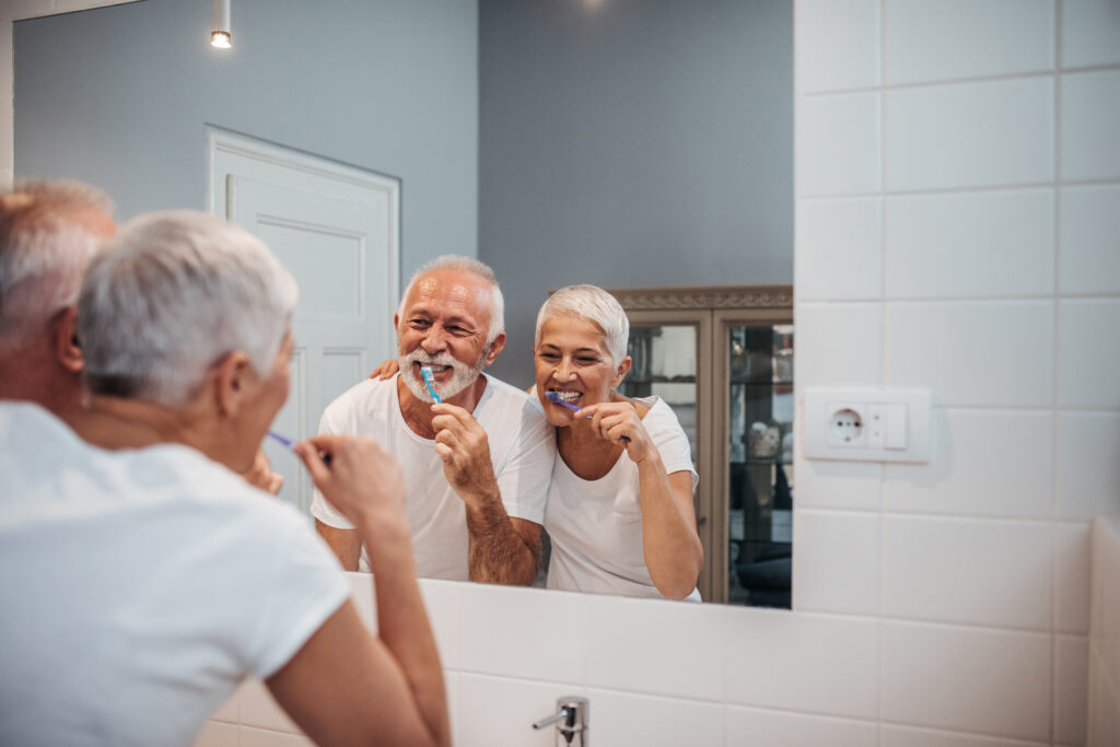 Oudere man en vrouw lachen terwijl ze samen voor de spiegel tandenpoetsen in een lichte, moderne badkamer