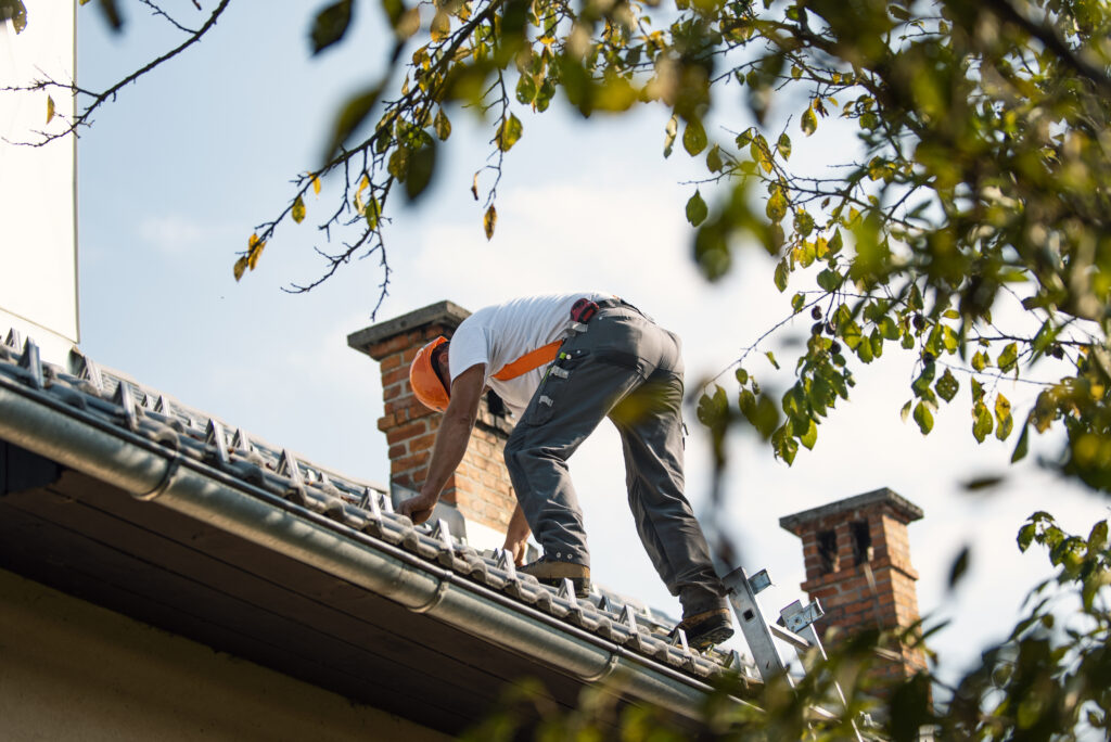 Man in werkkleding en helm werkt op het dak van een woning met behulp van een ladder, gefotografeerd van onderaf tussen bladeren