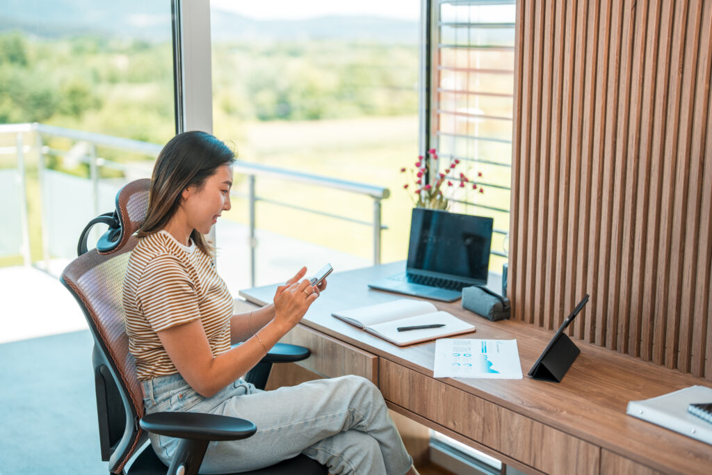 Vrouw zit in een lichte thuiswerkplek met laptop, telefoon en ergonomische bureaustoel.